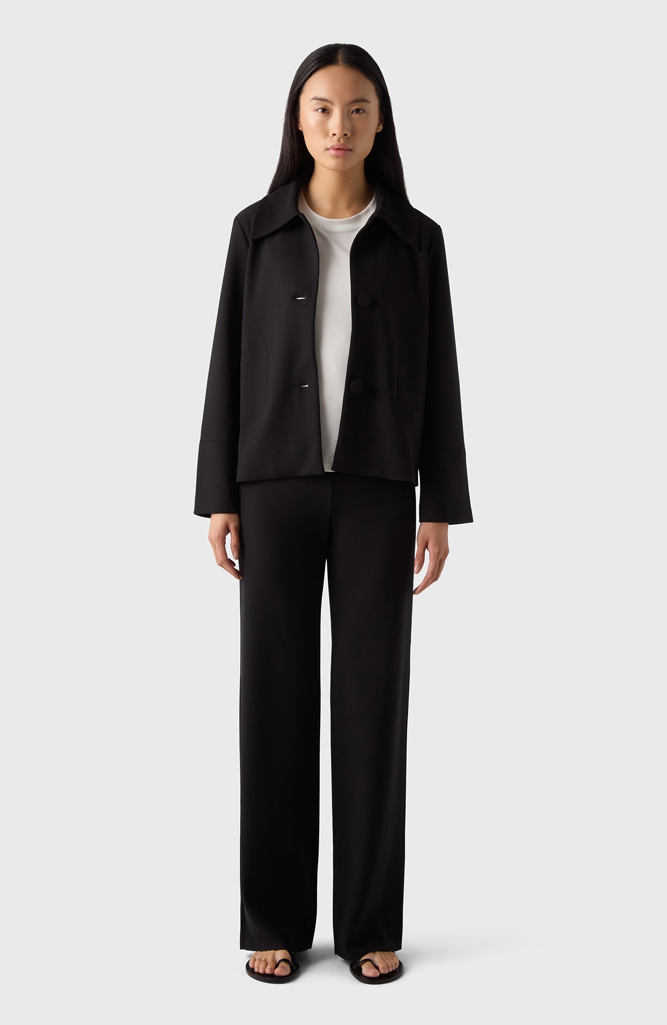 A woman standing, wearing black trousers and a black jacket with a V-neckline.