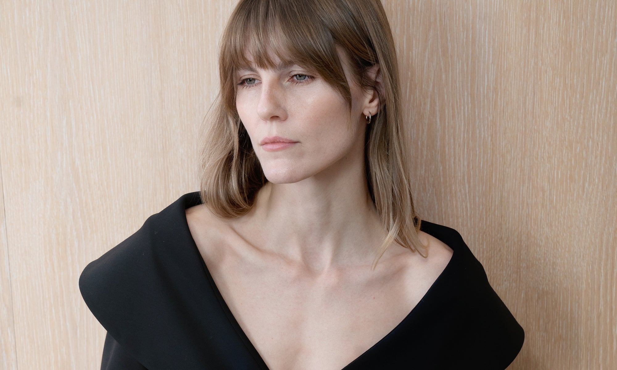 Woman in green jacket and black trousers leaning against a stone wall.